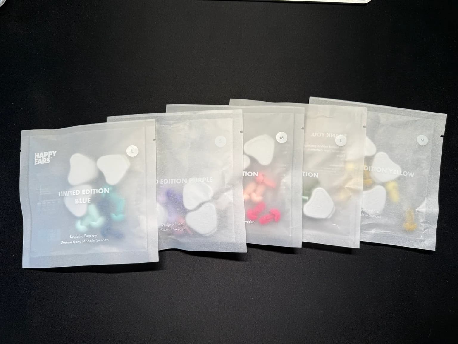 Fem förpackningar med Happy Ears öronproppar i olika färger, bland annat blå, lila, rosa, grön och gul, på ett mörkt bord.