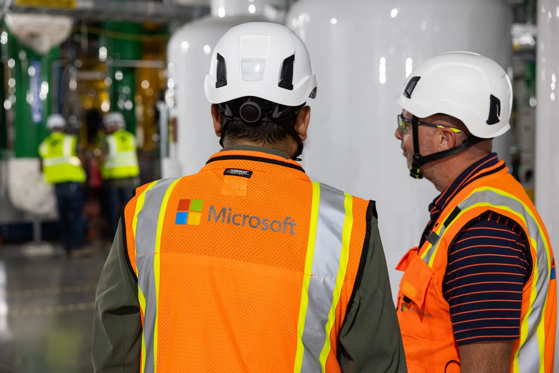 Två personer med vita skyddshjälmar och orange reflexvästar i en datacentermiljö. Microsofts logotyp syns på den ena västen.