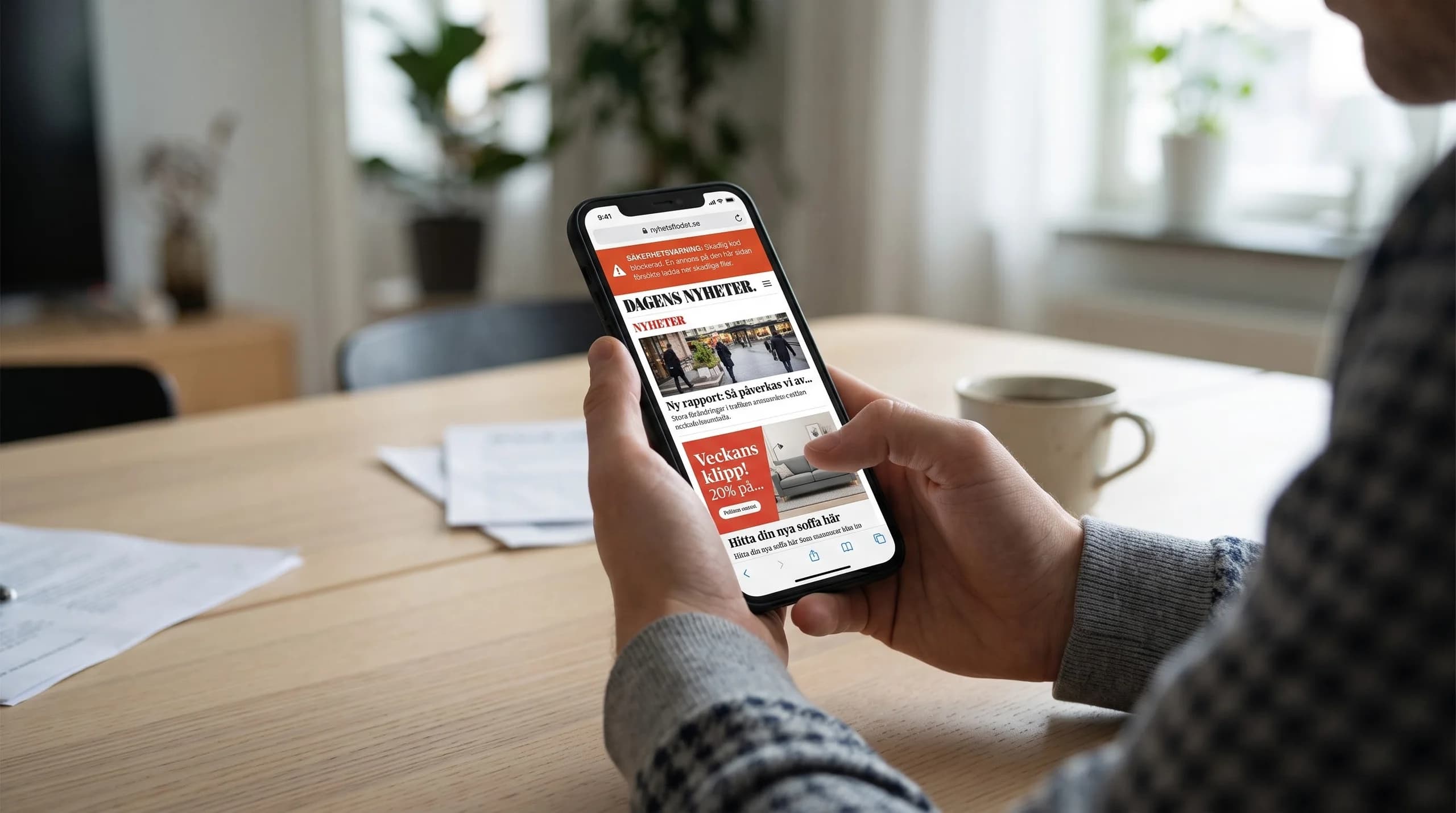 En person sitter vid ett ljust träbord och håller en mobil som visar en nyhetssida med annons och en röd säkerhetsvarning.