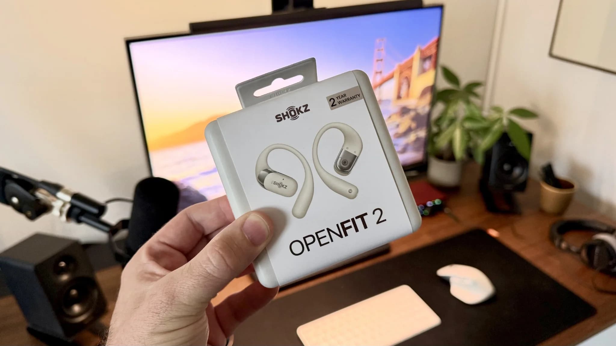 Förpackningen till Shokz OpenFit 2 hålls upp framför ett skrivbord med datorskärm.