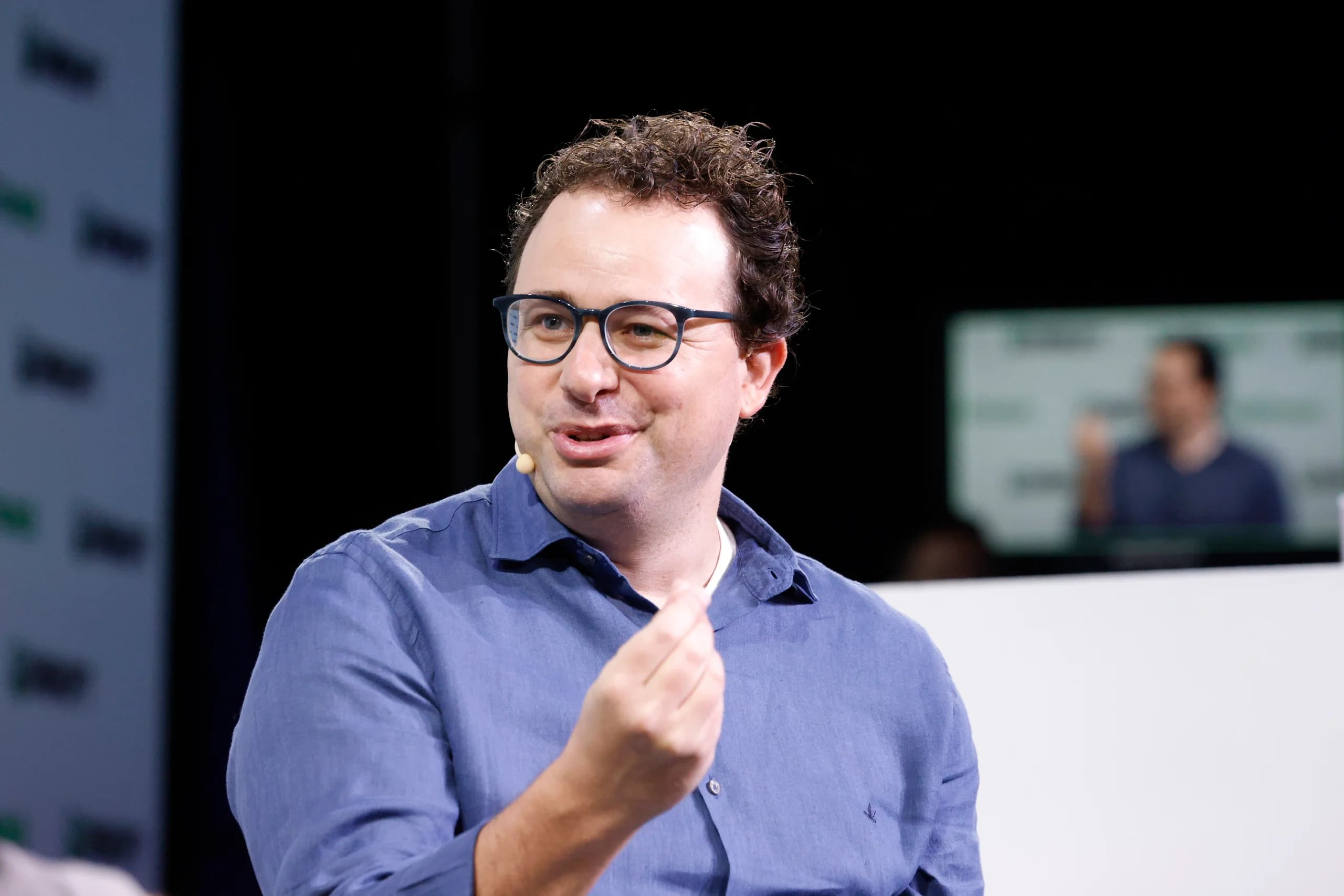 En man med glasögon, blå skjorta och headsetmikrofon gestikulerar under ett scensamtal vid TechCrunch Disrupt 2023.
