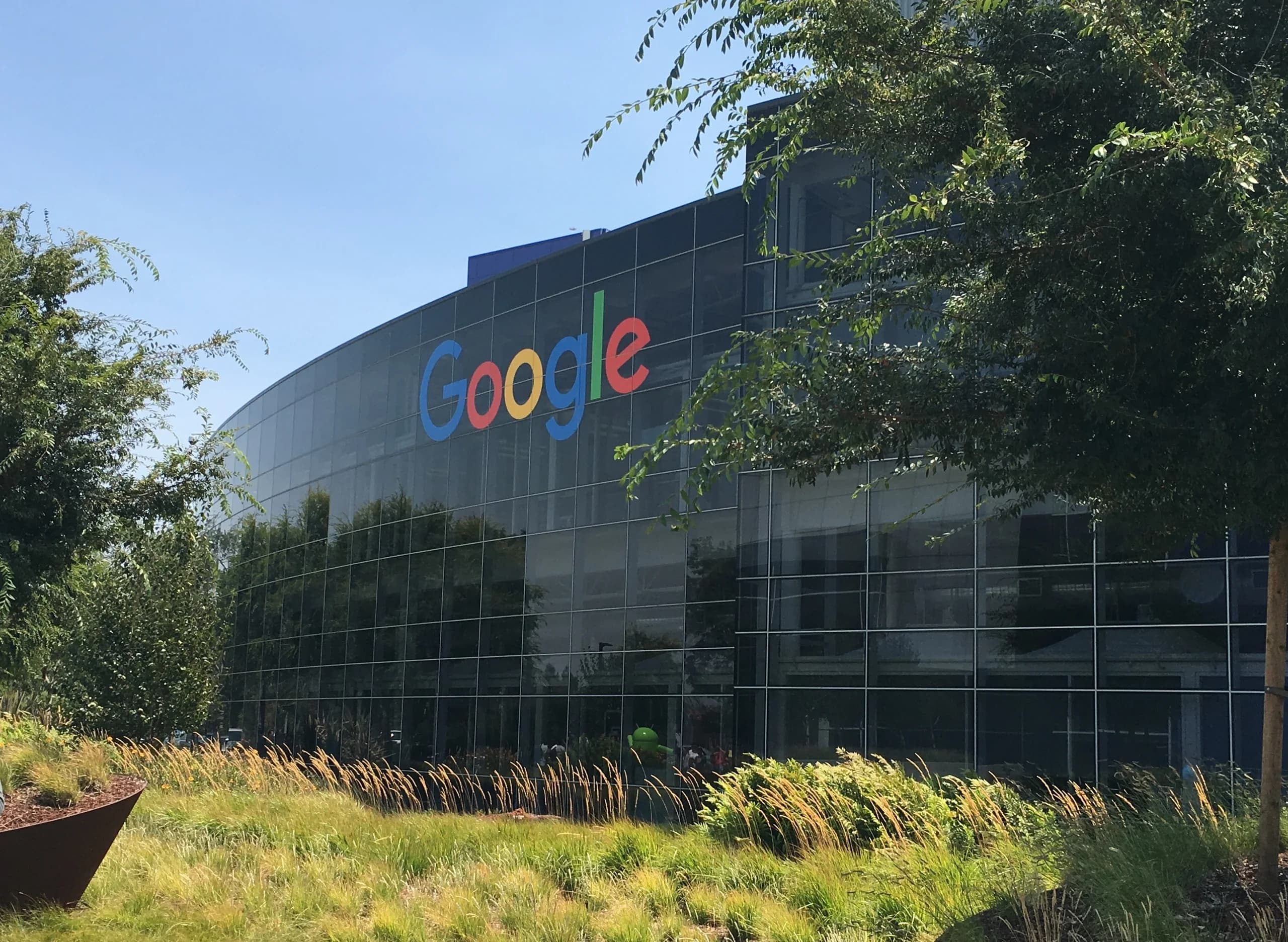 Googles logotyp på glasfasaden till Googleplex i Mountain View, med träd och gräs i förgrunden.