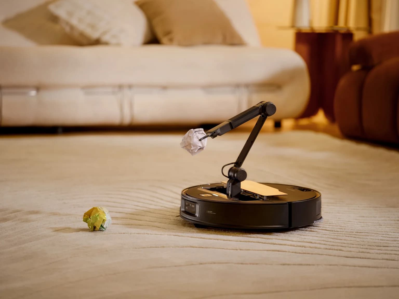 En robotdammsugare med en utfälld robotarm plockar upp en ihopknycklad pappersboll från en matta i ett vardagsrum.
